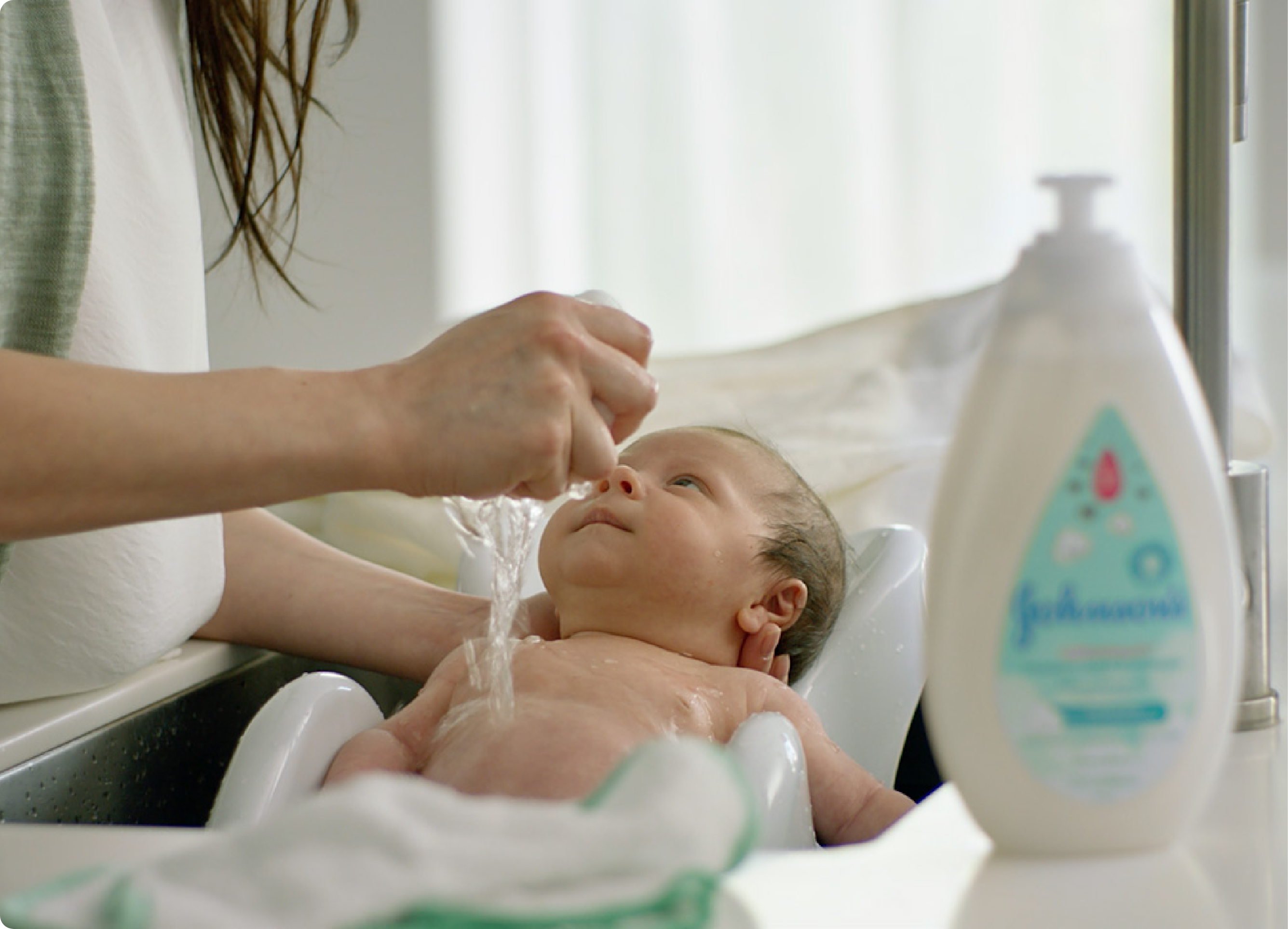 Baby being bathed in a sink, with their mother pouring water over the baby's head. A bottle of Johnson's Baby shampoo is placed on the right side of the image.
