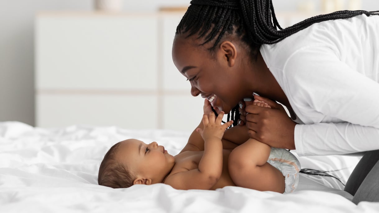 Madre sonriente jugando con su bebé en una cama blanca, mostrando unión y felicidad.