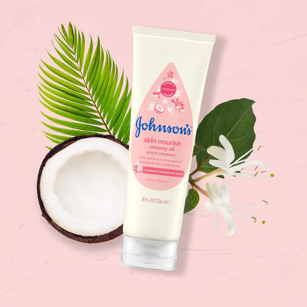 A tube of Johnson's skin nourish creamy oil lies near a halved coconut and green leaves against a pink background. Text on the tube reads: "Coconut & honeysuckle scents. 8 FL OZ (236 mL)."
