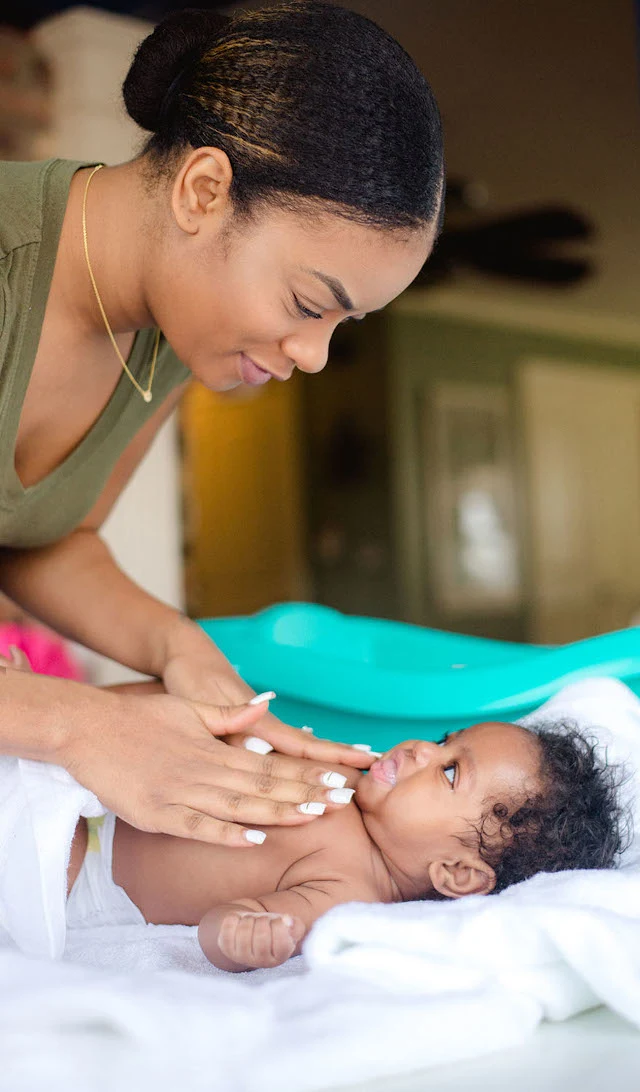 A woman tenderly touches a smiling baby lying on a white towel in a home setting with soft lighting and green and white furnishings in the background.