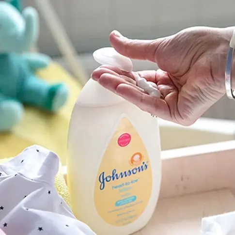 A hand dispensing Johnson's head-to-toe lotion from a bottle, with a baby’s clothes and toys in the background.