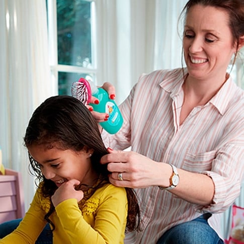 Mother using Johnson's baby No More Tangles Detangling Spray and combing daughter's wet hair, both smiling in a bright room.