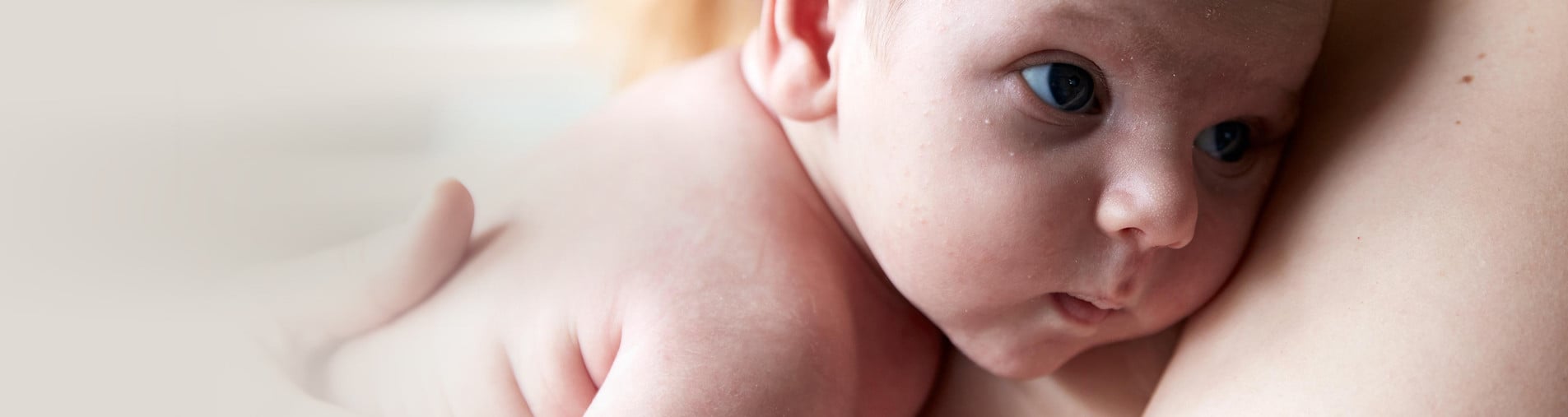 A baby rests its head on an adult's shoulder, gazing intently to the side. The background is softly blurred, drawing focus to the baby's face.