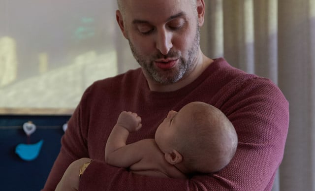 A bald man in a red knit sweater cradles an infant, looking down affectionately; the background shows a blue wall with a heart ornament and dim lighting.