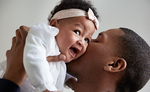 A father holding and playfully kissing his smiling baby girl who is wearing a white outfit and a headband with a bow.