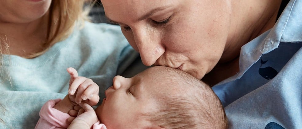 An adult holds a sleeping baby while gently kissing its forehead. Another person is visible in the background. The setting appears to be indoors and intimate.