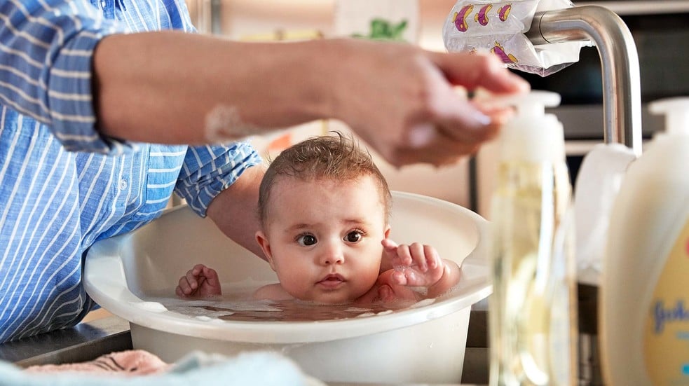 Bebé en una bañera, sostenido por un adulto que viste una camisa a rayas azules, en una cocina con jabón y grifo visibles cerca.