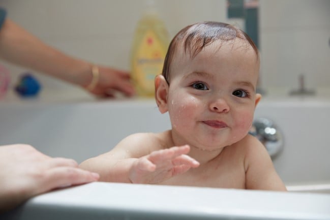 A baby with wet hair smiles while sitting in a bathtub, with a hand supporting them and a bottle of Johnson's baby shampoo in the background.