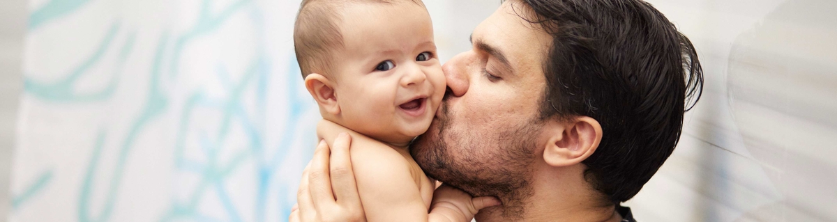 A father kissing his baby who is being held in his arms, both with joyful expressions.