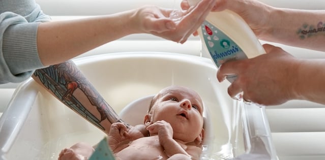 A baby is being bathed in a small tub by two people, one pouring liquid from a bottle labeled "johnson's" and the other holding the baby's head, in a well-lit room.