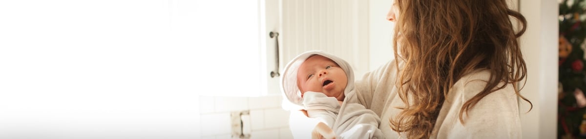 A person with long, wavy hair holds a newborn dressed in white attire. The baby looks up while being cradled. They are in a brightly lit room, with white tile and a window in the background.