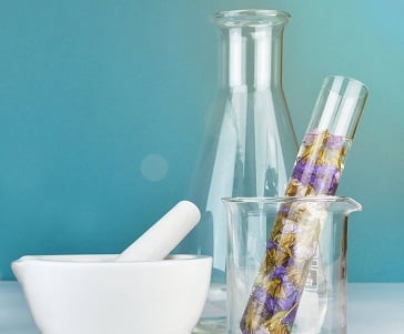 A mortar and pestle sit beside a beaker holding a test tube filled with dried flowers, with a glass flask in the background, all set against a teal backdrop.