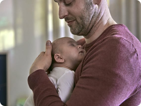 A man in a maroon sweater cradles a sleeping baby against his chest in a softly lit room with curtains in the background.