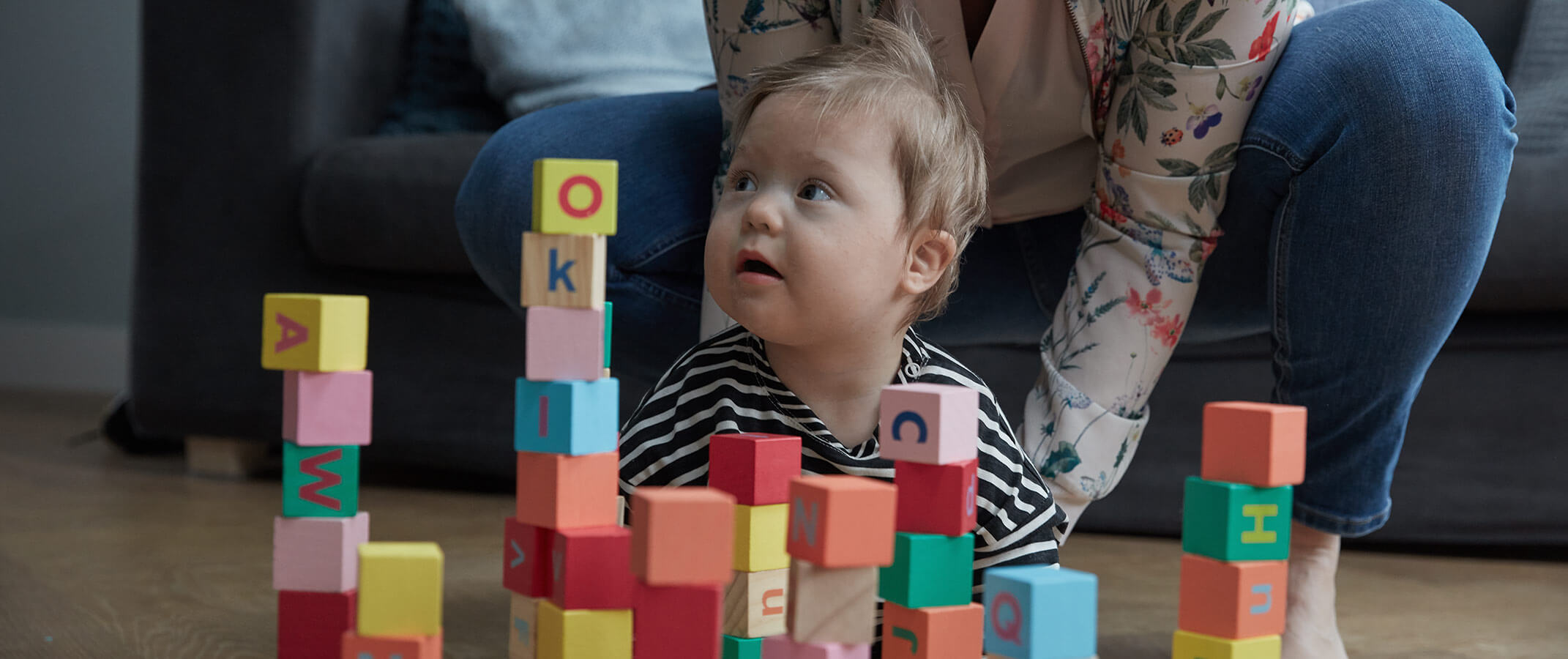 A toddler playing with colorful alphabet blocks, with his mother assisting in the background.