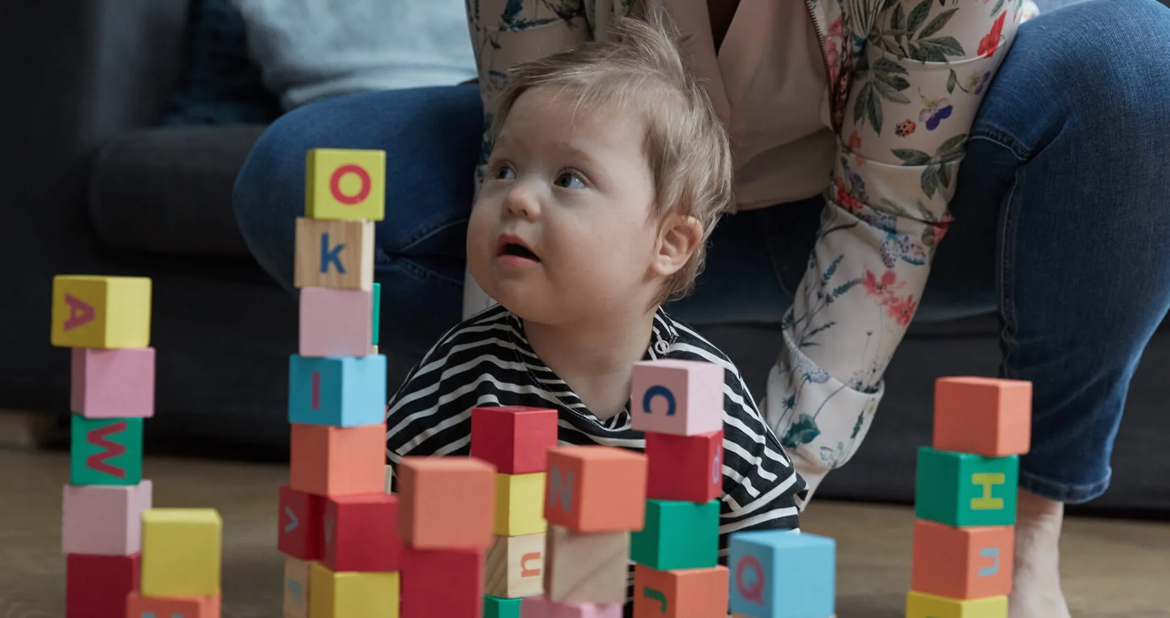 A toddler playing with colorful alphabet blocks, with his mother assisting in the background.