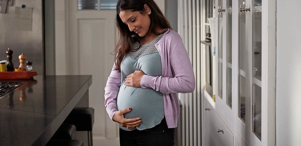 Pregnant woman cradles her belly, smiling and standing in a modern kitchen with white cabinets and a black countertop, creating a warm, domestic ambiance.