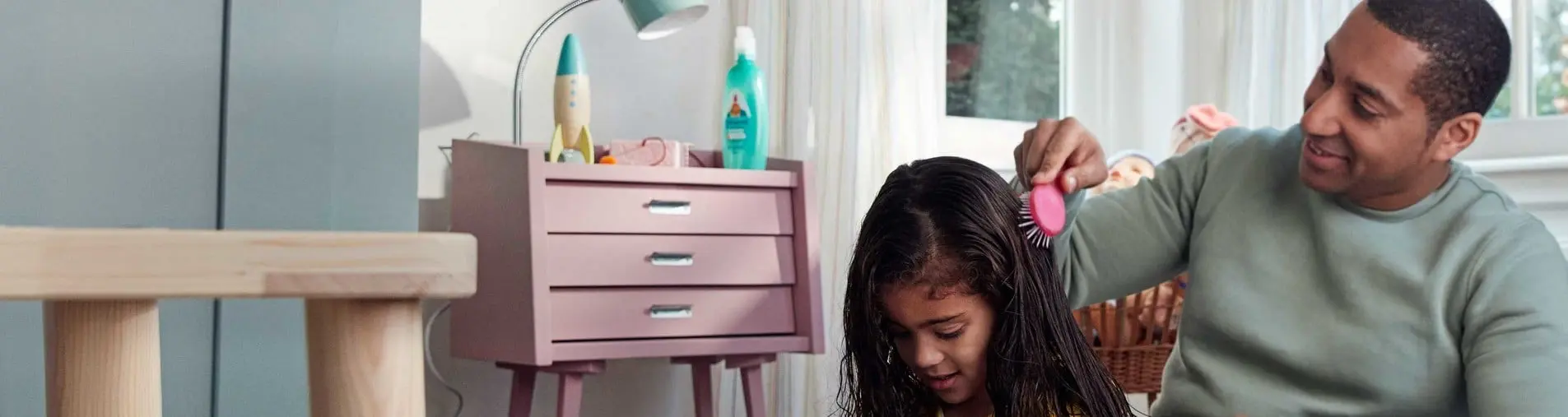 A smiling man brushes the wet hair of a seated girl in a bright room with a pink dresser and window in the background.