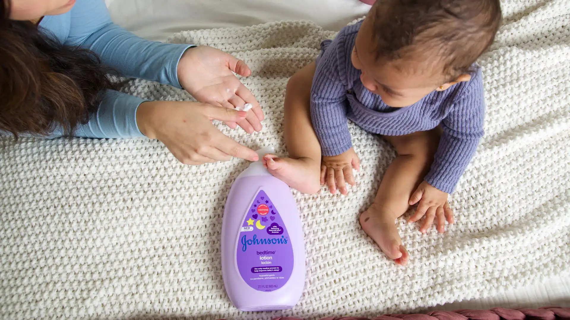 A baby wearing a purple outfit sits on a white blanket, while their mother applies Johnson's baby lotion to the baby's foot. The lotion bottle is placed in front of them on the blanket.