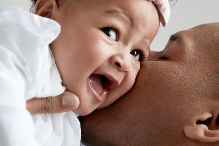 A close-up of a smiling baby being kissed on the cheek by a parent, capturing a joyful moment of affection.