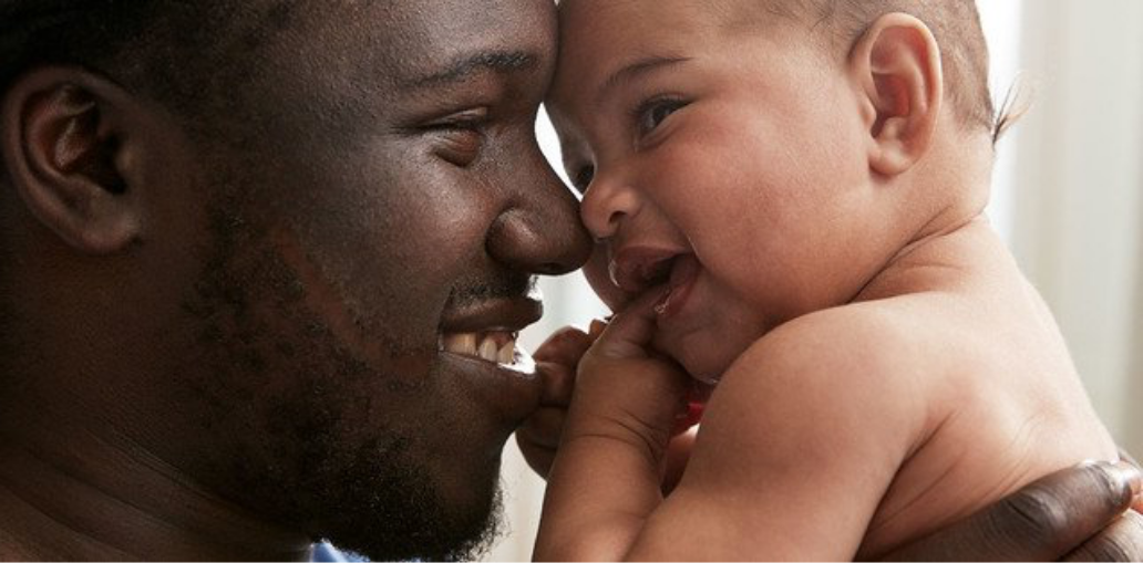 A smiling father holding and touching noses with his happy baby, both sharing a close and joyful moment.