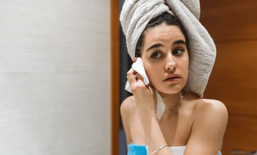 A person with a towel wrapped around their head, wearing a towel, wipes their face with a cotton pad in a bathroom with wooden and tiled walls.