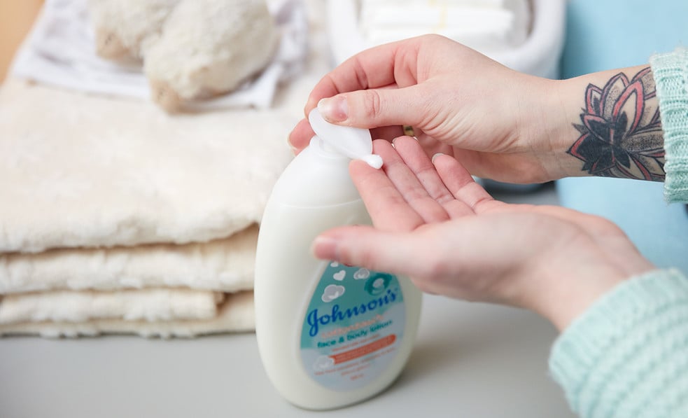 Hands pressing lotion from a Johnson's bottle onto a palm, with folded towels and baby items on a table in a bright, clean setting.