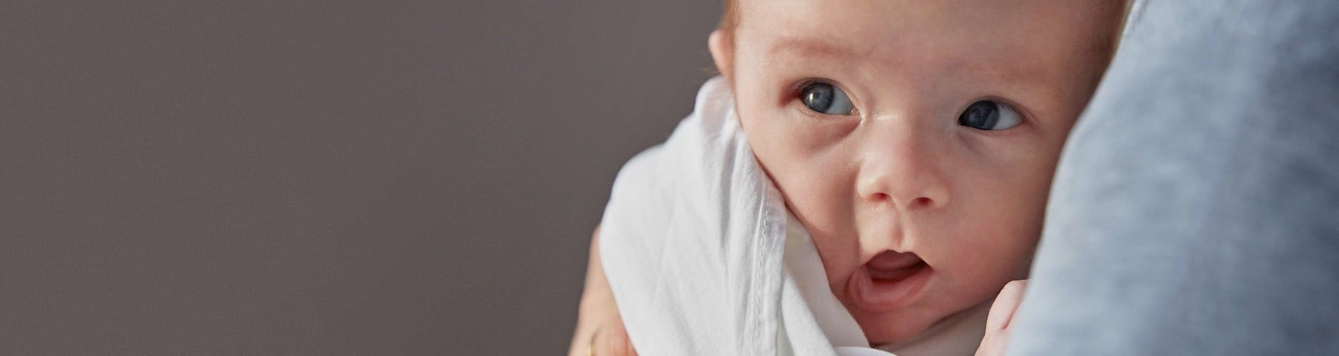 A baby wrapped in a white blanket is held in someone's arms, gazing curiously to the side with a neutral grey background.