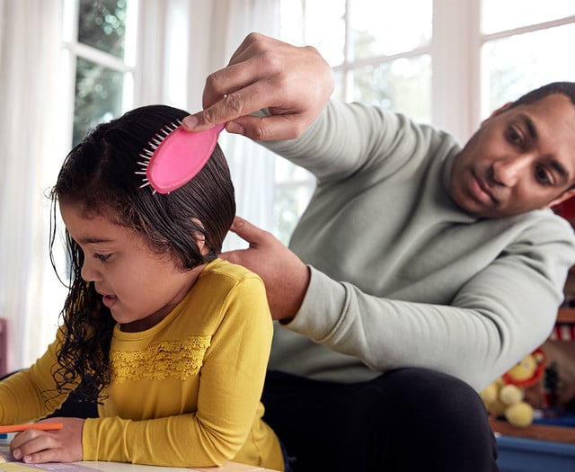 Un adulto cepilla el cabello mojado de un niño con un pincel rosa mientras el niño colorea en una mesa en una habitación luminosa con grandes ventanales en el fondo.