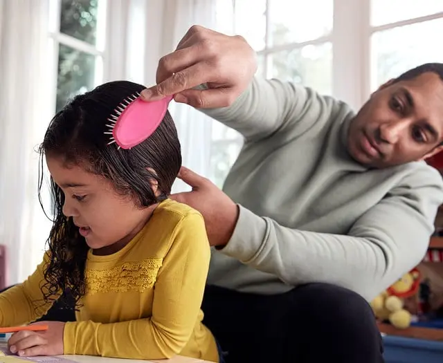 An adult brushes a child's wet hair with a pink brush while the child colors at a table in a bright room with large windows in the background.