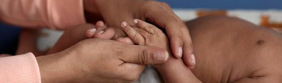 Adult hands gently holding a baby's hands; context includes a close-up of the baby's torso laying on a blanket.