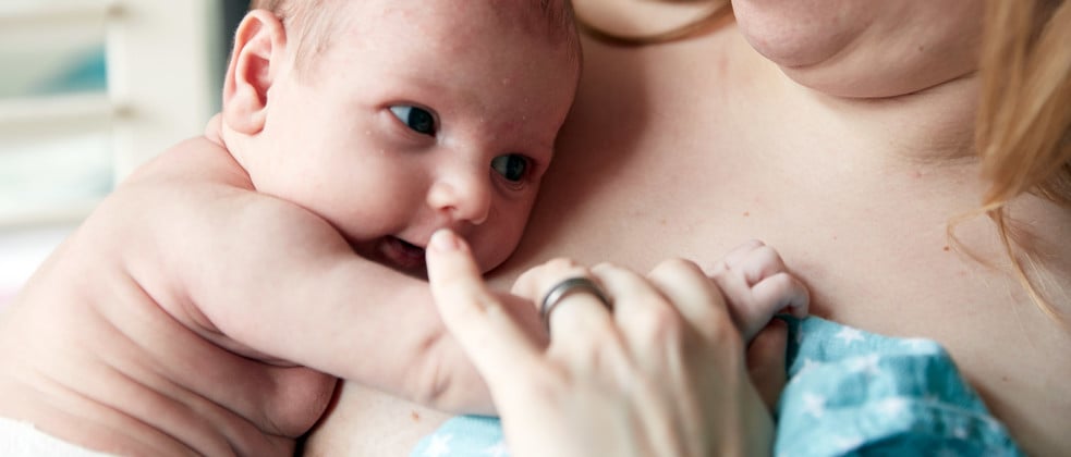 A newborn rests against a woman's chest, gently holding her finger, while she supports him with a soft blue blanket. The background features a window with partially closed blinds.