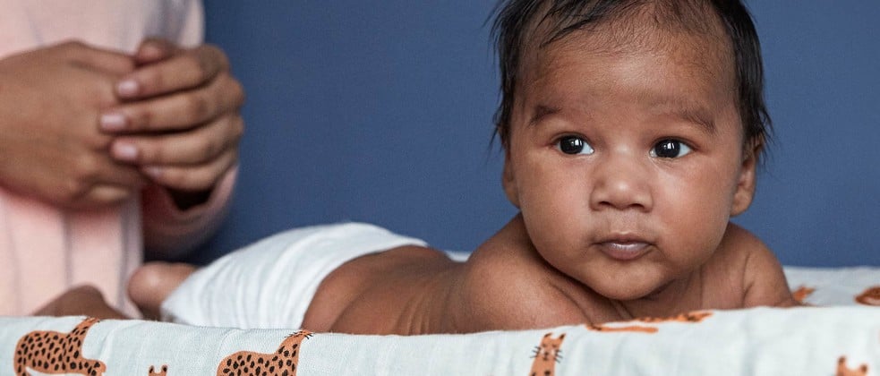 A baby lies on its stomach on a changing pad with a cheetah print, while an adult's hands clasp nearby against a blue background.