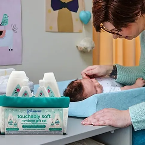 Parent caring for baby on changing table with Johnson's touchably soft newborn gift set in view.