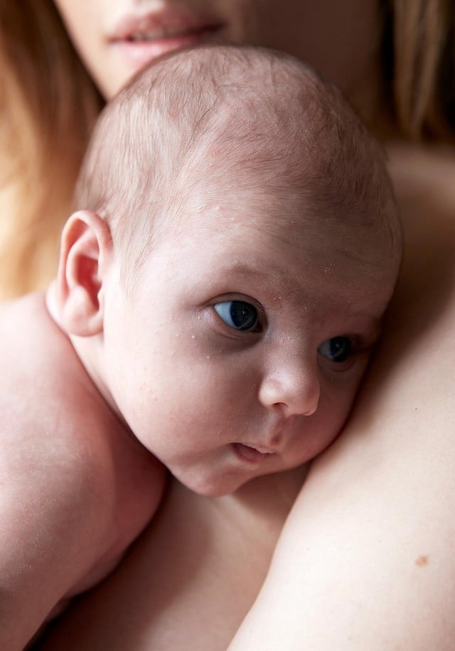 A newborn baby with large eyes rests against an adult's shoulder, closely cuddled, in a warm, intimate setting. The adult's face is partially visible in the background.