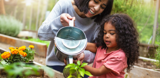 Woman and young girl watering plants in a garden, both smiling and enjoying the activity.