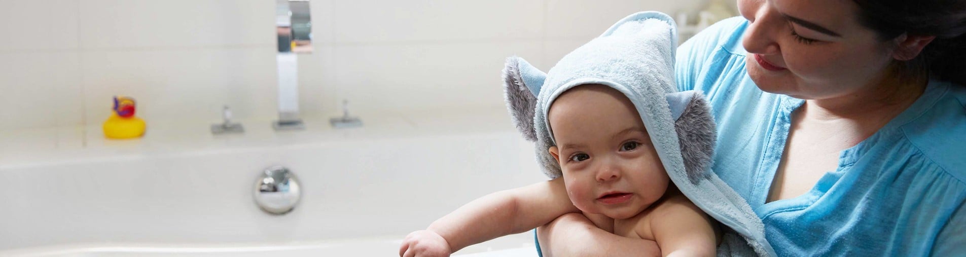 A baby, wrapped in an elephant-themed towel, sits in an adult's arms next to a bathtub with a yellow rubber duck and a silver faucet visible in the background.