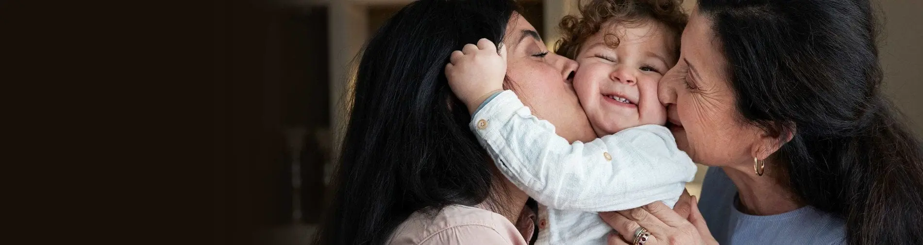 A mother and grandmother both kissing a smiling toddler on the cheeks, capturing a joyful family moment.