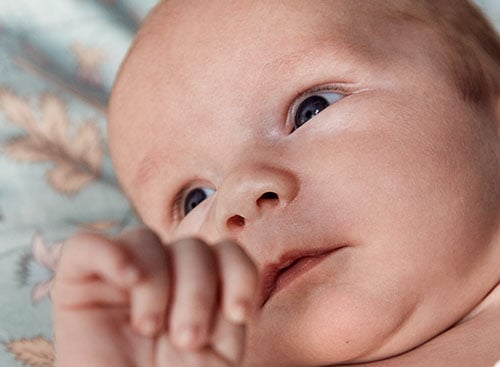 Close-up of a newborn baby with blue eyes and light skin looking upwards while lying on a patterned blanket.