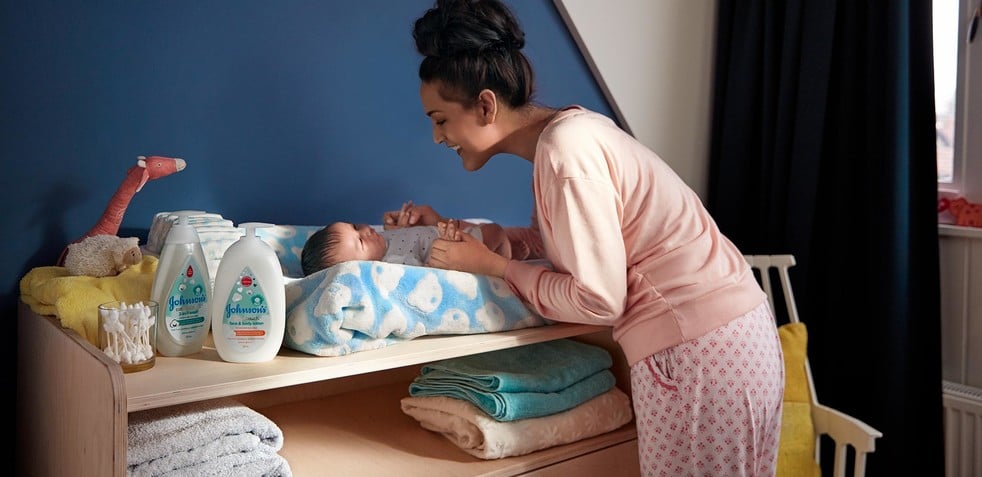 A woman in pink pajamas leans over a changing table, tending to a baby wrapped in a blue blanket, surrounded by Johnson's baby products, towels, and a stuffed giraffe.