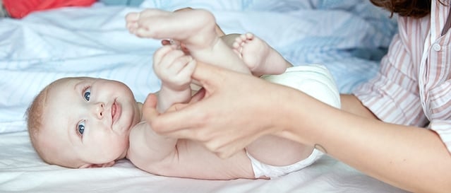 A baby lying on a bed is gently held by an adult, who is playing with the baby's feet. Bedding is visible in the background.