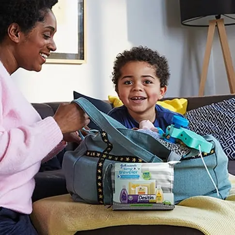 Mother and child packing a bag with a Johnson's Tiny Traveler Baby Gift Set, including four baby products and a free travel clutch.