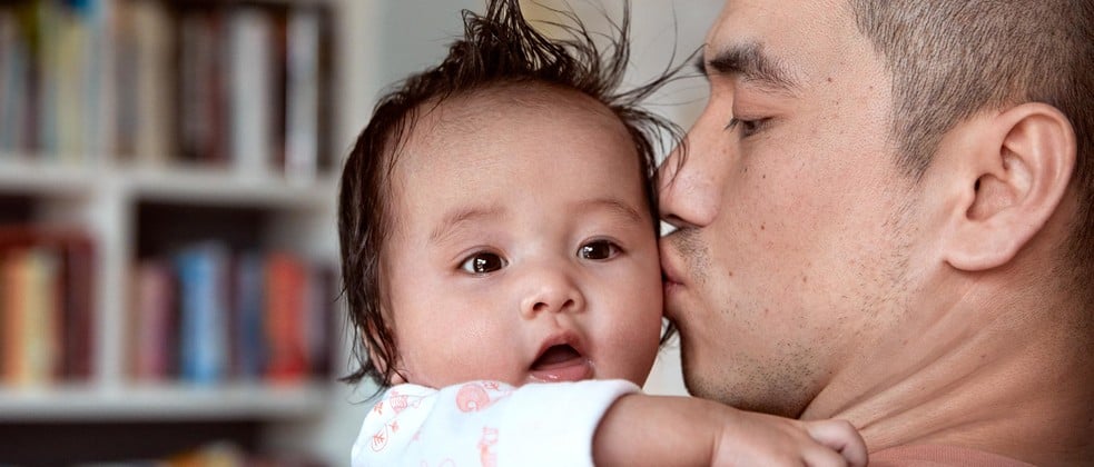 An adult tenderly kisses the cheek of a baby, who looks surprised. They are indoors with bookshelves blurred in the background.