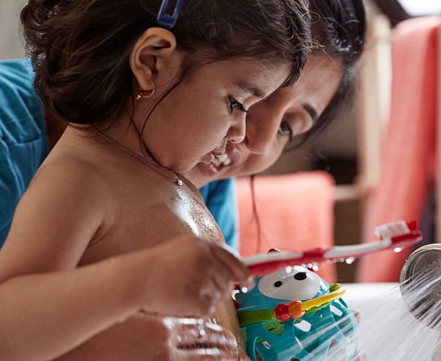 A toddler with a hair clip washes a toy, while an adult closely assists, in a home bathroom with orange towels in the background.