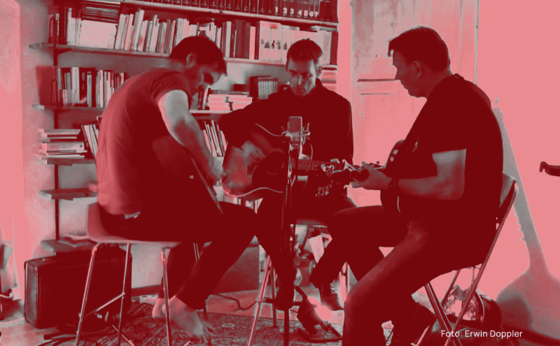 Christoph, Christian and Andreas sitting in a living room with a book shelf in the background and playing guitar.