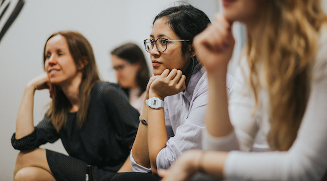 A photograph of a row of women side by side, looking forward and resting their chins in their hands in engaged listening postures. The appear to be audience members watching an event or class. 