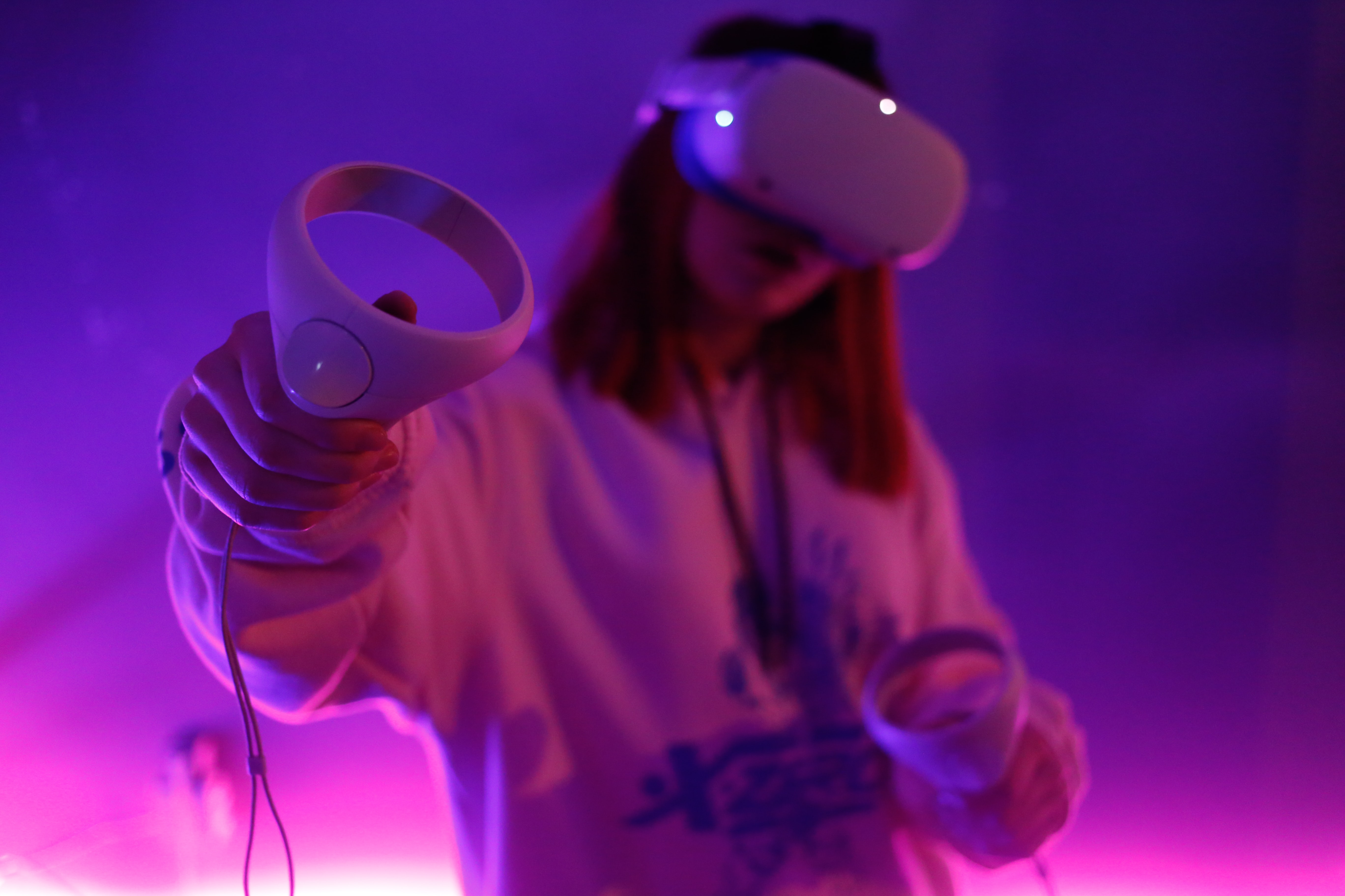 A photograph of a woman in a virtual reality room lit by purple light. She is holding a video gaming controller in her hand and wearing a consol on her face. She has shoulder-length brown hair and is wearing a white hoodie. 