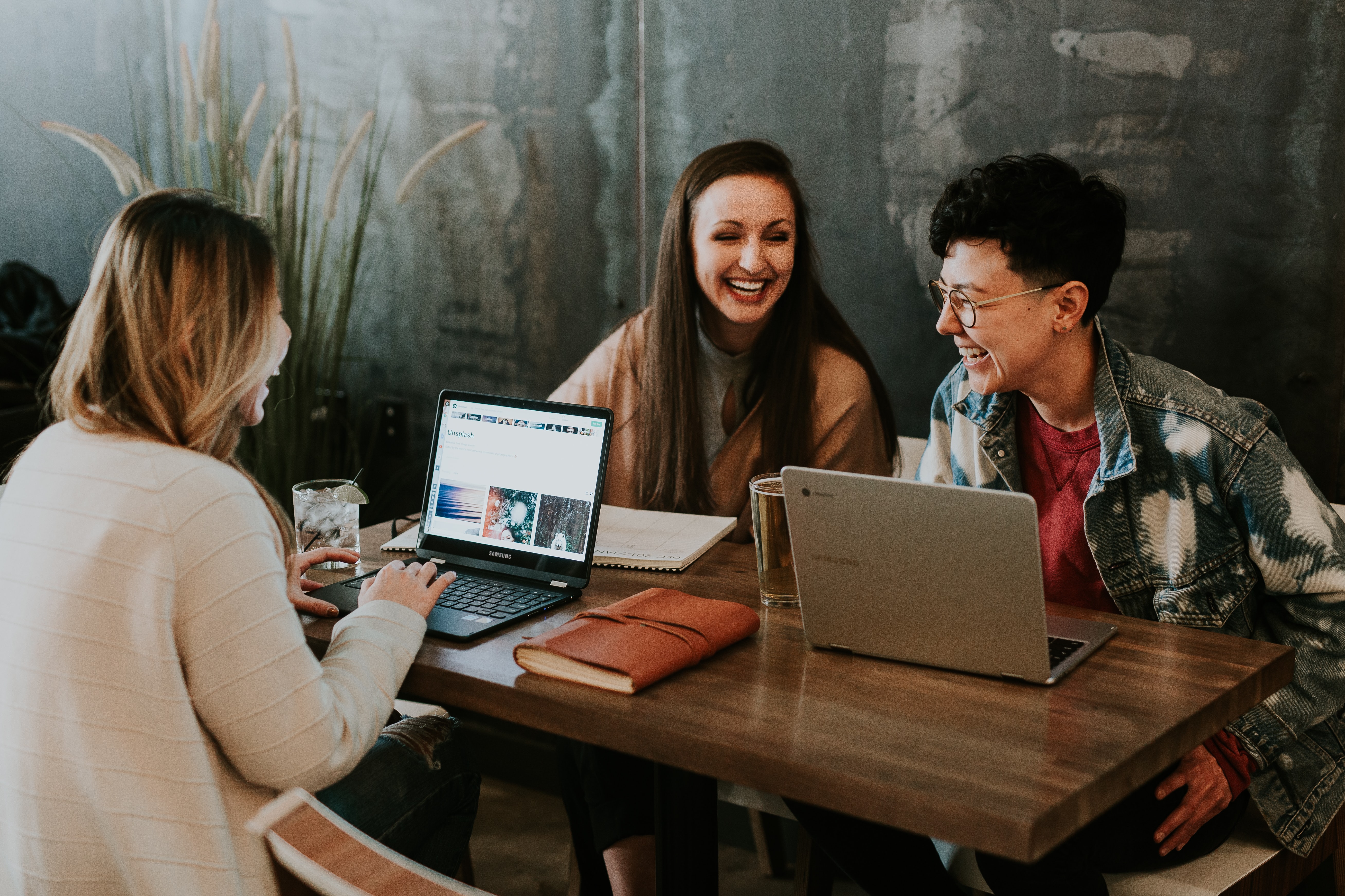 A photograph of three women sitting together at a wooden desk. On one side of the desk, two women are smiling joyfully, looking at a silver Mac laptop. Behind them is a backdrop of a grey wall. Beside them, a green-leafed plant can be seen just out of frame. One of the women has an A4 white notebook in front of her. Opposite them is another woman dressed in a white cardigan also smiling while looking at a silver Mac laptop. Next to her is a leatherbound journal.  