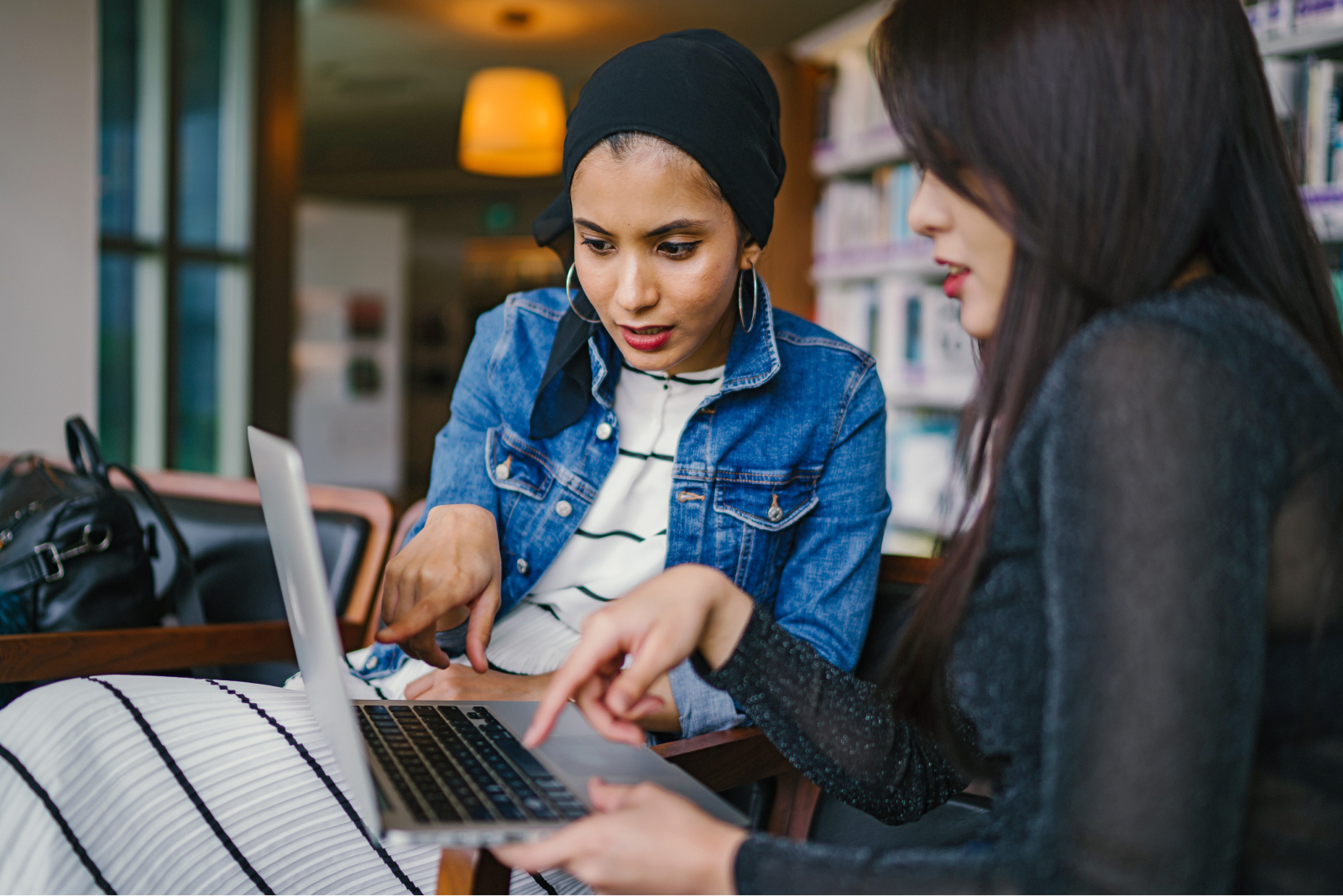 A photograph of two young women engaged in conversation while pointing at a silver Mac laptop. One of the women is wearing a striped black and white dress, a blue denim jacket, gold hoop earrings, and a black scarf covering her hair. The woman next to her has long black hair and is wearing a black sweater. In the background is a lit yellow ceiling light and a set of bookshelves.