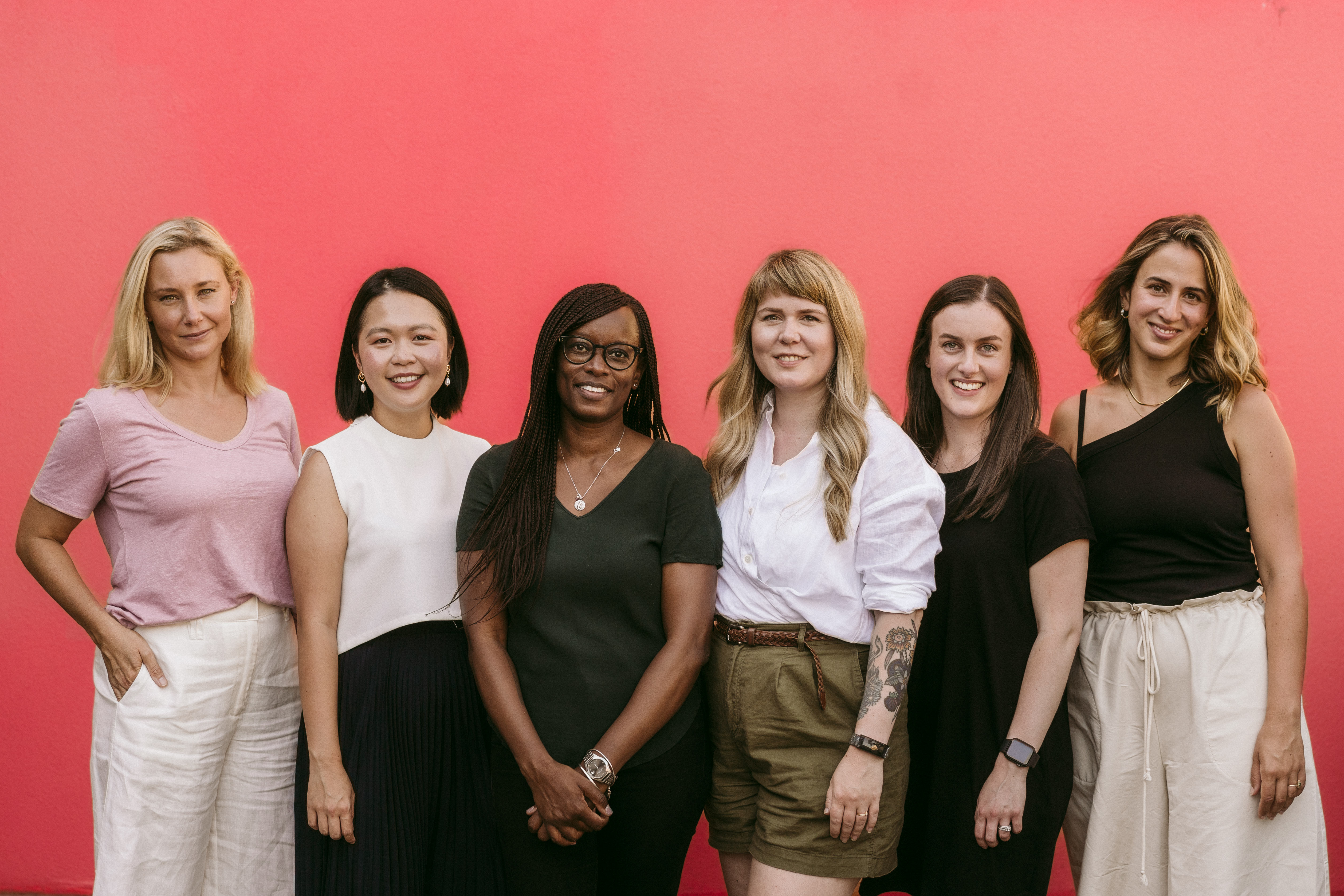 A photograph of a group of 6 women standing against a backdrop of a red wall, looking directly at the camera while smiling.