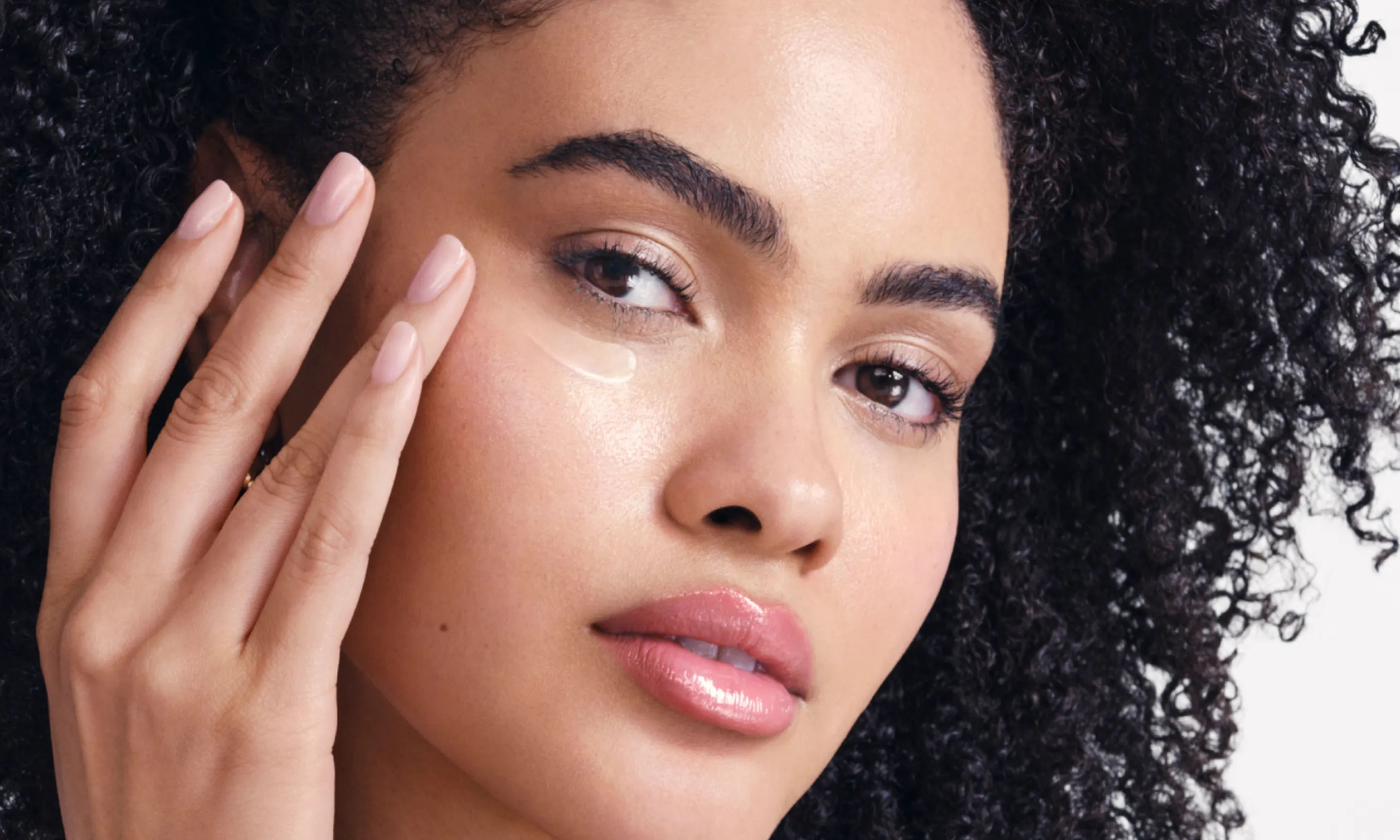 A headshot of a young woman with curly brown hair, applying a product to her under-eye area with her finger.
